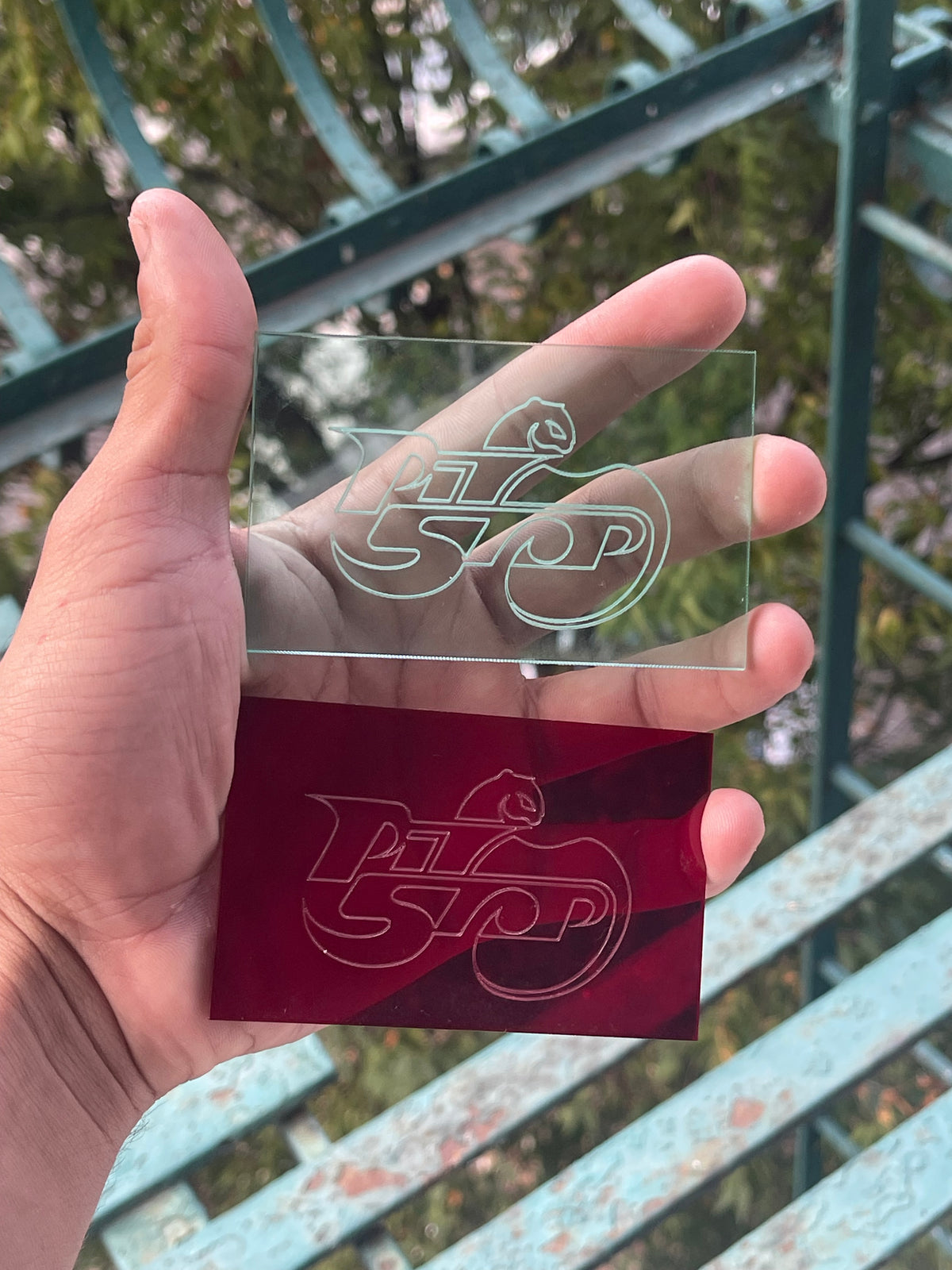 Hand holding two laser-engraved acrylic cards—one mint/ transparent and one red—displaying the PIT//STOP logo, demonstrating custom etched branding and merchandise prototypes, against a blurred outdoor background